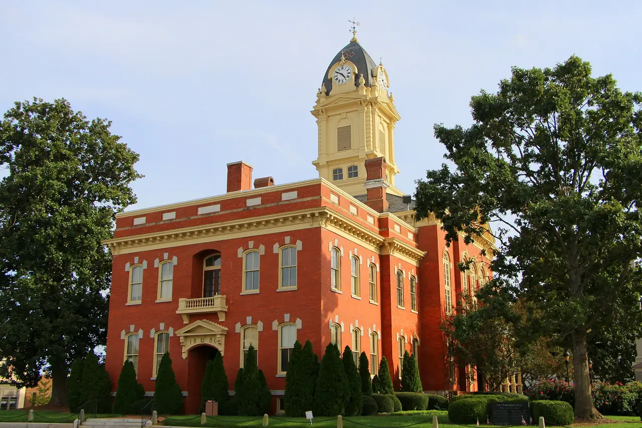 The red-brick Union County Courthouse in downtown Monroe, North Carolina, with its cream cupola and clock above a row of evergreen shrubs and two large shade trees